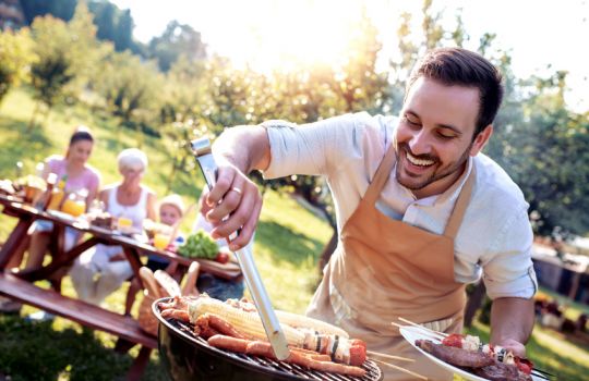 Fröhlicher Mann grillt Fleisch, Gemüsespieße und Maiskolben auf einem Holzkohlengrill. 