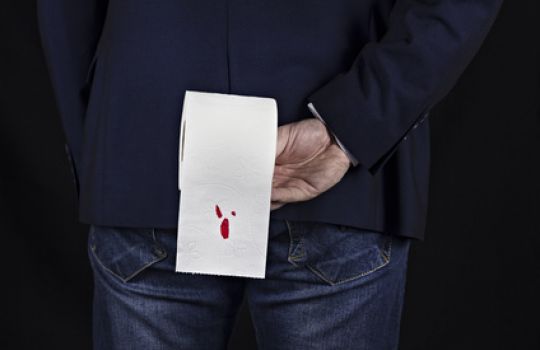 Mann hält Klopapierrolle hinter dem Rücken, auf der hellrotes Blut zu sehen ist.