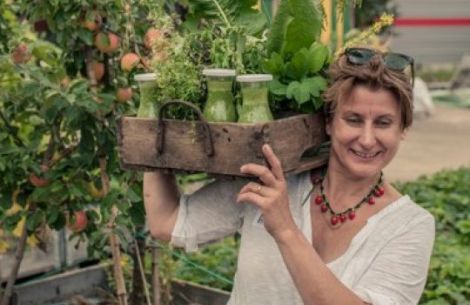 Gabriele Bräutigam trägt eine Holzkiste, in der sich Pflanzen und Glasflaschen mit grünen Smoothies befinden.