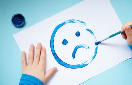 Kinderhände malen einen blauen, traurigen Smiley auf ein Blatt Papier