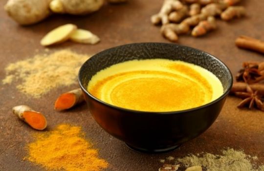 Ein Schälchen, gefüllt mit goldener Milch, im Hintergrund die Gewürze.