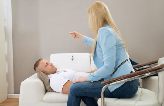 Eine Frau sitzt mit einem Pendel vor einem Mann, der auf dem Sofa liegt und die Augen geschlossen hat.