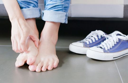 Fotografie des Fußbereichs einer Frau, die sich am rechten Fußrücken kratzt, daneben Turnschuhe