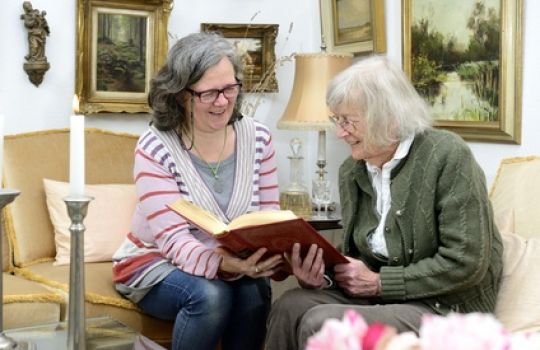 Freundin und ältere Dame lesen gemeinsam in einem Buch als Demenzprophylaxe.