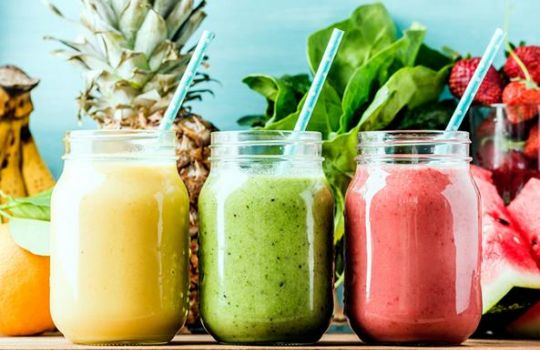 Gelbe, grüne und rote Smoothies im Glas. Dahinter Bananen, Ananas, Kopfsalat und Erdbeeren.